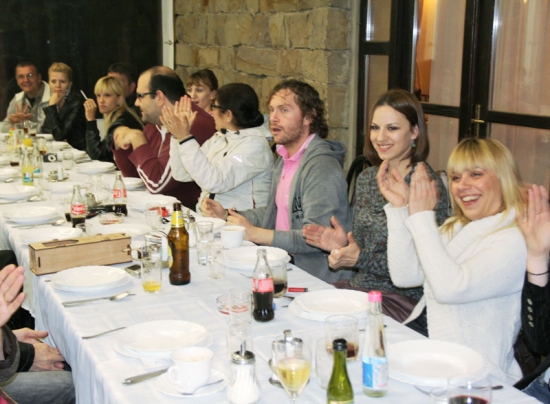 Participants sit at a long table, smiling and clapping their hands during dinner at Novi Kneževac. 