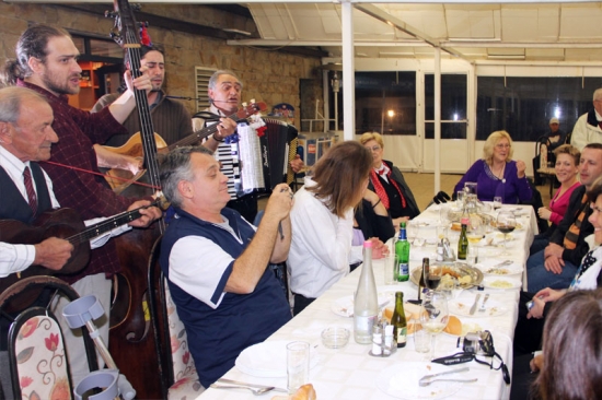 Four men play musical instruments on one side of a long table while participants smile and watch on the other side.