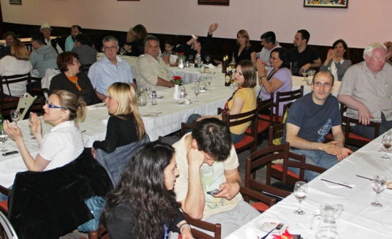 Participants are seated beside long tables with  white tablecoths, wine glasses and water pitchers. Some are clapping.