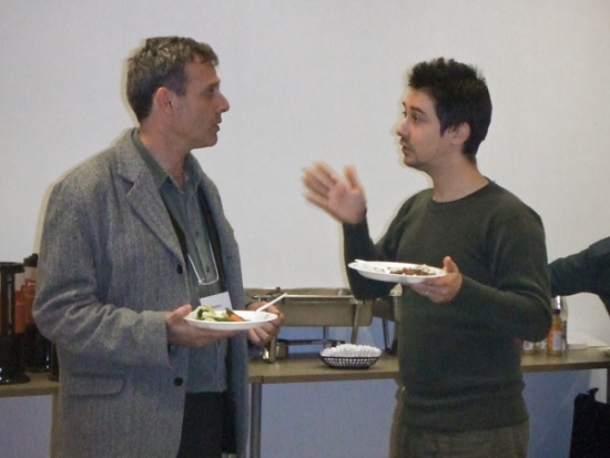 Two participants have a conversation over a bite to eat. Lunch is served in the background as the two participants stand to have their discussion. 