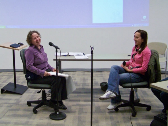 Melanie and Iphigenia are seated facing each other with a microphone in front of Melanie. They are both smiling and laughing.