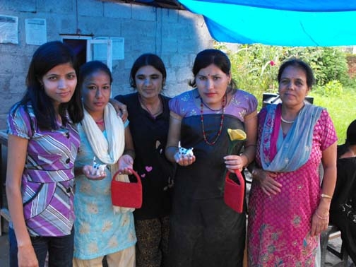 <p>Five women from stand outside beneath a blue tarp with crafts they created.</p>
