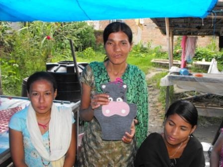 <p>Two women seated and a third woman standing in the middle holding a knitted hippo craft.</p>
