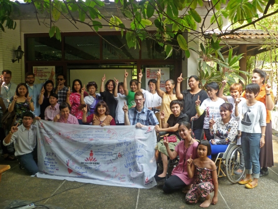 <p>People pose around a large white banner full of writing and drawings. Most people are holding up a single finger in the air.</p>
