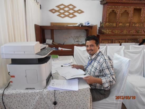 Kumar  Regmi sitting at a desk smiling at the camera working on participants lists for the country training in Nepal.  The photo is taken from his left side and he is turning his head to look at the camera.