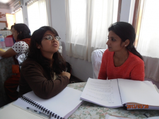 Two participants are seated at a table with their books open in front of them. In the background there is another participant with her back turned. 