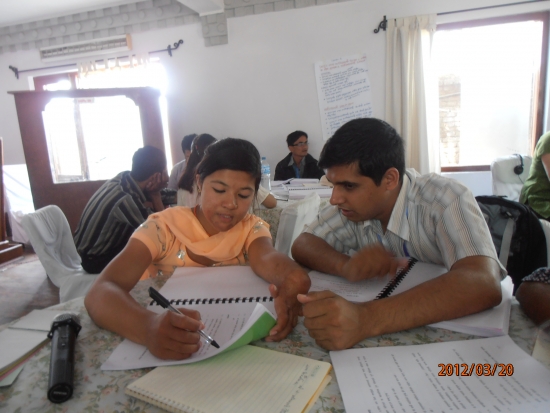 <p>
	Two participants are sitting inside the training room as they point to a page in their training manuals with other training items on their table including note books and a microphone.</p>
