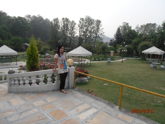 Chona stands on a patio section in front of the garden outside of the hotel. In the background there are covered seating areas, trees and grass with a path running through the middle.