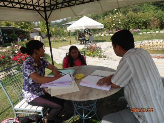 <p>
	A table outside in the garden, with three people seated at the table. There are training materials on the table and all three people are facing each other.</p>
