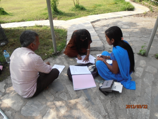 Three participants sit on the ground as the one in the centre of the photo writes and the other two (left and right in the photo) look on. 