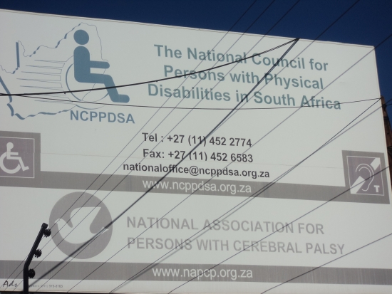 Large white sign with blue lettering saying: National Council for Persons with Physical Disabilities in South Africa. Also on the sign are the phone number, fax and email of the organization. 