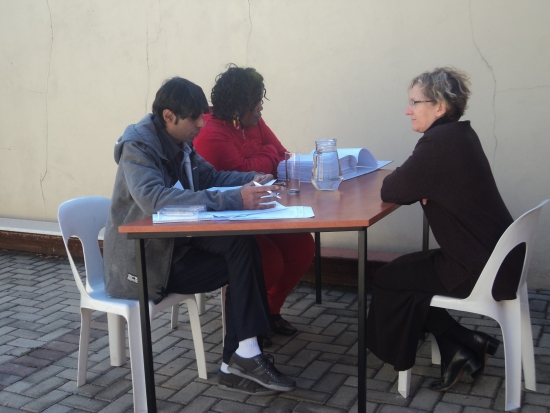 <p>
	Helene Combrinck is sitting at a table being interviewed face to face by Imtiaz Moola and Zama Ngwenya.</p>
