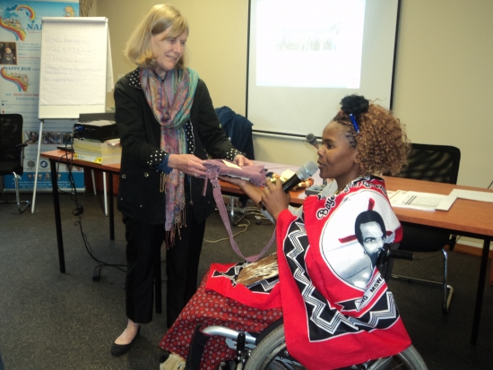 Prof. Marcia is standing and receiving a gift from Bongiwe Malope, inside the training venue. In the background there is a flip chart, tables and chairs. 