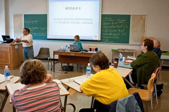 Steve Estey is speaking near the front of the room while participants sit at small desks and listen.