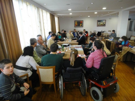 Milenko is speaking to everyone from Montenegro in a country meeting. Everyone is gathered around a set of tables that fills the back of the training room. 