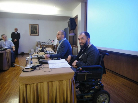 Zoltan and Samir are seated behind a table at the front of the training room , there is a large presentation screen behind them. 