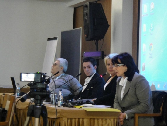 Presenters sit behind a table in the training room. They speak into microphones in front of them. A camera is visible in front of them. 