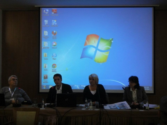 Milenko  and Rados are seated beside Deputy of Minister of Labor and Social Care, Ms. Remzija Ademovic and PR Manager of Mayor of city of Budva, as they give their opening remarks. There is a large presentation screen behind them. 