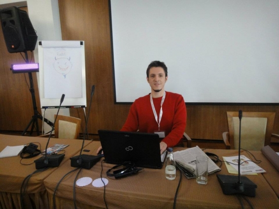 Rados sits down at the front of the room and smiles for the camera. Behind him is the screen and in front of him are the microphones and an open computer.