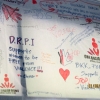 <p>A close up of a white cloth banner covered with writing. One prominent slogan by Chona states &quot;DRPI supports women to be free of violence!&quot;. Most other slogan call to stop violence against women.</p>

