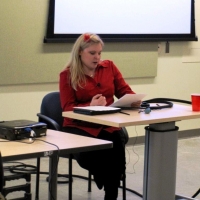 A woman sits and reads from a paper at a desk at the front of the room.