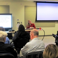 <p>
	A woman sits at a desk while presenting at the front of a large room. Participants sit facing her in the foreground.</p>
