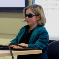 A close-up photo of a woman with sunglasses presenting while sitting at a desk.