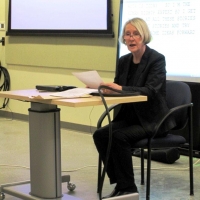 A woman presents while sitting at a desk at the front of a large room.
