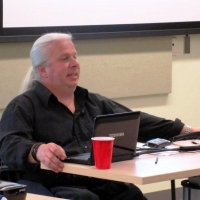 A man sits behind a desk at the front of the room and presents.