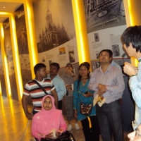 Bangladeshi and Nepali team listen to a tour guide in a long hallway filled with photographs and information on Thailand.