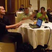 Christopher Lytle, Lauro Purcil and Rannil Sorongon sit at a table with a laptop. Participants sit at tables in the background. 