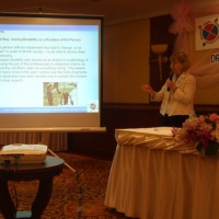 Marcia Rioux, stands beside a slide presentation and speaks into a microphone. 