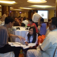 Khamphon, Phoutsady and Lapkeo sit together and have a discussion. There are people and tables in the background. 