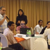 Mr. Harka Bahador stands next to a table with seated participants and speaks into a microphone. 