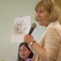 Marcia Rioux holds up a pencil sketch of herself while speaking into a microphone. Chona Sabo looks on.