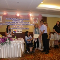 Lapkeo Somchanmavong smiles and holds his certificate with Saowalak Thonkuay and Marcia Rioux while other facilitators look on. 