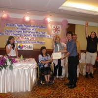 Roong-aran Wongkaewmoon smiles while holding his certificate with Saowalak Thonkuay. Marcia Rioux and Nathan Bond have both their hands raised in the air and smile while other facilitators look on. 