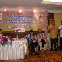 Lauro Purcil stands and speaks into a microphone while other facilitators sit and listen in the background. 