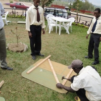 A man kneels to hammer together two boards outside while three other men stand around watching.