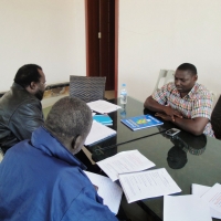 <p>
	Three men sit at a large shiny black table covered with training materials.</p>
