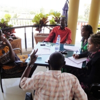 Mr. Ted, Nkatha Murungi, Mercy Mugure, Moses Mayanja and Suzan Kirima sit and talk around a table on a veranda.