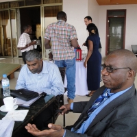 Mr. Robert Mkozho and Jace Nair sit at a table on a veranda. Other participants are in the background standing near a table with coffee carafes.
