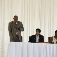 A man in a suit stands and speaks into a microphone with two others seated to the right