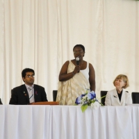 Honourable Minister Jeanne d'Arc Mujawamaliya stands and speaks into a microphone while others sit to her left and right.