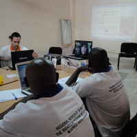 Severin Rwamucyo and Oswald Tuyizere look at a laptop with Paula Pinto's face on screen. Chris is also seen working on a laptop