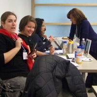 Three women are sitting in a row at a table crowded with food and papers.