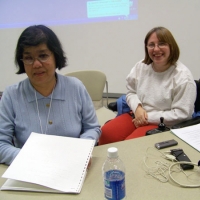 Two participants smile for the photo sit side by side for a photo. On the desk is translation equipment and a bottle of water. 
