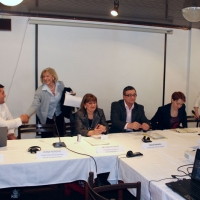 <p>
	Representatives are seated at a long rectangular table adorned with a white tablecloth, microphones and headsets.</p>
