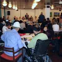 Mr. Mihali stands at the front of the room and speaks into a microphone while training participants sit around tables listening.