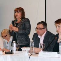 Ms. Petrušić  stands and speaks into a microphone with other organizational representatives seated on either side.